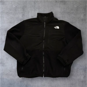 Svart fleecejacka från The North Face - Svart fleecejacka från The North Face med hög krage och hel dragkedja framtill. Jackan har två sidofickor med dragkedja och en bröstficka. Materialet är mjuk fleece och polyester, perfekt för kyliga dagar. Logotypen är broderad i vitt på bröstet och ryggen. Går att zippa in i vissa North Face jackor.