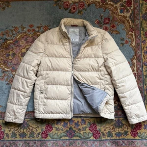 Brunello Cucinelli Jacka - Snygg beige pufferjacka från Brunello Cucinelli med grått foder och diskreta detaljer. Jackan har dragkedja, knappar och två sidofickor. Tillverkad i Italien med lätt och mjukt material som ger en clean och modern look.