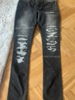 Grå slitna jeans  - Säljer ett par svarta jeans med coola slitningar och råa detaljer på framsidan. Jeansen har raka ben och klassisk femficksdesign. Materialet är jeans och färgen är mörkgrå/svart med blekta partier och tydliga trasiga effekter på låren och knäna.