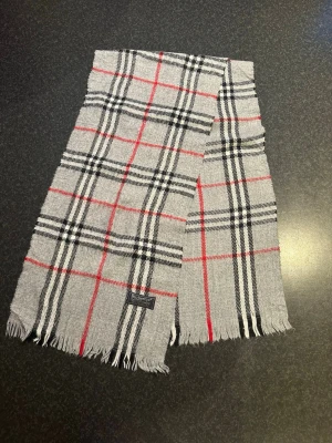 Grå rutig Burberrys kashmir - Säljer en grå väst från Burberrys i mjuk kashmir med klassiskt rutigt mönster i svart, vitt och rött. Västen har fransad nederkant och en snygg Burberrys-label. Perfekt för dig som gillar tidlös design och lyxigt material.