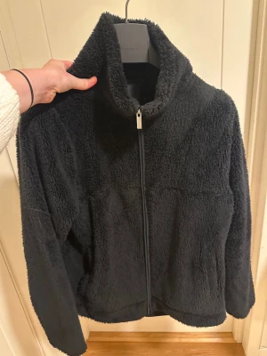 Svart teddyjacka från Lager 157 - Mjuk och fluffig svart teddyjacka från Lager 157, modell Cosy. Jackan har hel dragkedja framtill, hög krage och långa ärmar. Tillverkad i 100% polyester för extra mysig känsla. Perfekt för kyliga dagar när du vill vara både bekväm och stilren.