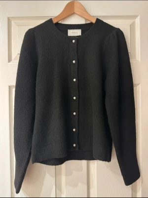 Svart kofta med pärlknappar från Neo Noir stl 38 - Snygg svart kofta från Neo Noir med rund hals och långa ärmar. Koftan har pärlknappar framtill och är stickad i en mjuk blandning av polyester, akryl, ull och elastan. Perfekt för dig som gillar stilrena och klassiska plagg med en twist. Endast provad så oanvänd stl 38