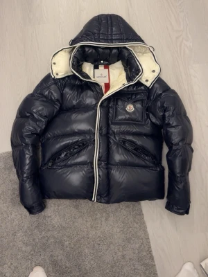 Moncler Branson navy pufferjacka  - Svart pufferjacka från Moncler med glansig finish, huva och vit insida med röd och marinblå rand. Jackan har dragkedja, fickor med dragkedja och Moncler-logga på bröstet. Storlek 3 (passar bäst M storlek) användt ett par gånger, lika som ny