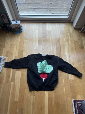 Svart sweatshirt med rädistryck Mini Rodini - Cool svart sweatshirt från Mini Rodini med ett stort rädistryck i grönt och rött framtill. Tröjan har rund hals, ribbade muddar vid ärmslut och nederkant samt långärmad passform. Materialet är mjuk bomull som är skön att ha på sig.