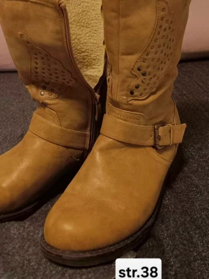 Bruna boots med spänne, storlek 38 - Säljer ett par snygga bruna boots i storlek 38 med dekorativa nitar och mönster på skaftet. Skorna har rund tå, låg klack och spänne vid ankeln. Tillverkade i mocka med grov gummisula som ger bra grepp. Perfekt för dig som gillar western-inspirerad stil.
