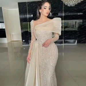 Champagnefärgad långklänning med pärlor - Säljer en elegant långklänning i champagnefärg med glittriga pärldetaljer och asymmetrisk axel. Klänningen har lång ärm på ena sidan och vackra draperingar samt transparenta partier. Perfekt för bal eller festligheter.