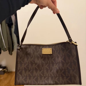 Brun axelväska Michael Kors - Snygg brun axelväska från Michael Kors med MK-logga över hela väskan. Väskan har guldfärgade metalldetaljer, dragkedja och ett mönstrat innerfoder. Perfekt storlek för det viktigaste och har flera kortfack inuti.