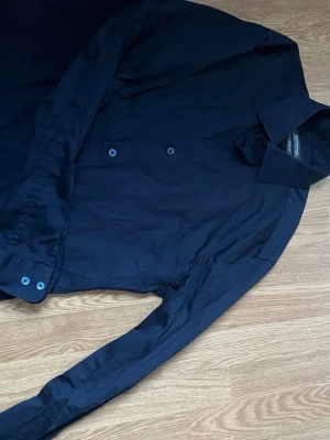Mörkblå skjorta Dressmann slim fit - Snygg mörkblå skjorta från Dressmann i slim fit-modell. Tillverkad i bomull med stretch för extra komfort. Klassisk krage och knappar framtill, långärmad och stilren design som funkar till många olika looks.