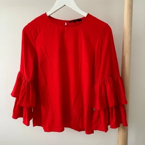 Röd volangblus från Vero Moda - Snygg röd blus från Vero Moda med volangärmar och rund halsringning. Blusen har långa ärmar med dubbla volanger.