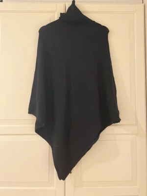 Svart polotröja poncho från Vila - Svart polotröja poncho från Vila med asymmetrisk form och hög krage. Tröjan är loose fit och har ett stilrent, minimalistiskt utseende. Materialet är stickat och passar perfekt för kyliga dagar. Enkel att slänga över en outfit för extra värme och stil.