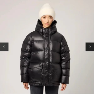 Svart pufferjacka från Singular Society  - Säljer en svart pufferjacka med huva och stora fickor framtill. Jackan har en glansig finish, dragkedja och knappar framtill samt snörning i huvan. Perfekt för kalla dagar och riktigt skön att bära tack vare den vadderade designen.