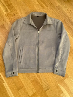 Grå mockajacka med dragkedja - Snygg grå mockajacka med klassisk krage och raka linjer. Jackan har två sidofickor, hel dragkedja framtill och svarta tryckknappar vid ärmsluten. Materialet är mjukt och ger en stilren look som funkar till många outfits.