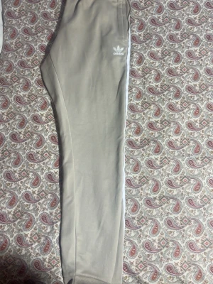 Beige mjukisbyxor från Adidas - Snygga beige mjukisbyxor från Adidas med klassiska vita ränder längs sidorna och Adidas-logga på höften. Byxorna har ribbade muddar vid bensluten och är gjorda i mjukt material som är skönt att ha på sig. Perfekta för chill eller träning.