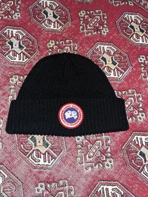  Canada Goose mössa  - Säljer en svart ribbstickad mössa från Canada Goose med klassisk logga framtill. Mössan är i mjukt material och har en bred uppvikt kant. Perfekt för kalla dagar och snygg till alla outfits.