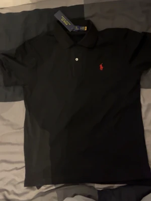 Svart pikétröja från Polo Ralph Lauren - Klassisk svart pikétröja från Polo Ralph Lauren i slim fit. Tröjan har korta ärmar, krage och två knappar framtill. På bröstet finns den ikoniska röda Polo-loggan. Tillverkad i mjuk bomull för en skön känsla. Skickar inom 24 timmsr