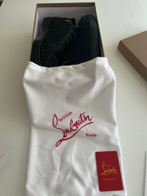 Christian Louboutin svarta sneakers med nitar - Svarta sneakers från Christian Louboutin med coola svarta nitar på tån och klassisk röd sula. Skorna har snörning och är tillverkade i mocka med en platt sula. Perfekt för dig som vill sticka ut med en lyxig och edgy stil. 