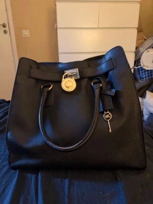 Svart handväska Michael Kors - Snygg svart handväska från Michael Kors med guldfärgade detaljer, lås och nyckel med MK-logga. Väskan har två handtag och en axelrem med kedja. Tillverkad i skinn med stilren och klassisk form, perfekt för dig som gillar lyxiga accessoarer.