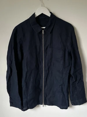 Mörkblå overshirt från J.Lindeberg - Snygg mörkblå overshirt/skjortjacka från J.Lindeberg i 100% linne. Jackan har klassisk krage, hel dragkedja framtill och knappdetaljer vid ärmslut. Perfekt att slänga på över en t-shirt för en chill och clean look. 5M18