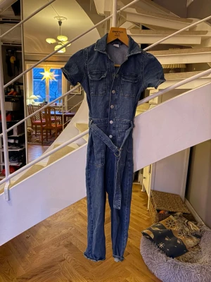 Blå jeans jumpsuit med bälte strl 134 - Snygg blå jeans jumpsuit med korta ärmar, knappar framtill och bälte i midjan. Flera fickor både upptill och nedtill ger en cool och praktisk look. 