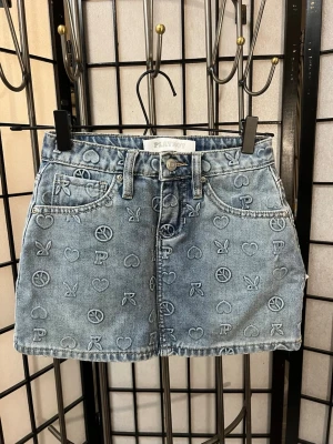 Blå jeanskjol med mönster från Playboy - Säljer en helt ny blå jeanskjol från Playboy med broderade symboler som hjärtan, peace-tecken och kaninloggan över hela kjolen. Kjolen har klassisk femficksdesign, knapp och dragkedja framtill samt bälteshällor. Perfekt för dig som gillar streetstyle och coola detaljer.