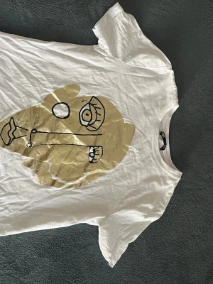 Vit t-shirt med guldtryck från Zara - Säljer en vit t-shirt från Zara i storlek S med ett stort abstrakt ansikte i guld och svart tryck på framsidan. T-shirten har rund hals och korta ärmar, tillverkad i mjuk bomull. Perfekt för dig som gillar unika prints och vill sticka ut.