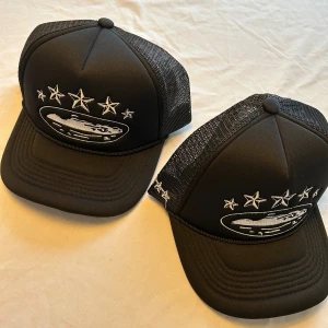 Svart Corteiz truckerkeps med stjärnor - Säljer en svart truckerkeps från Corteiz med vit broderad grafik framtill, inklusive fem stjärnor och en oval logga. Kepsen har mesh på sidorna och bak, samt justerbar passform. Perfekt för dig som vill ha streetwear-vibe och snygg detalj till outfiten.