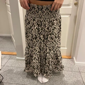 Mönstrad långkjol med volang - Säljer en svart och beige långkjol med snyggt grafiskt mönster och bred smock i midjan. Kjolen har volang nertill. Så snygg!