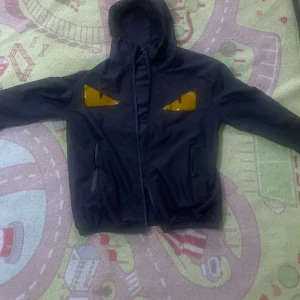 Svart vindjacka med gula detaljer - Svart vindjacka med huva och dragkedja framtill. Jackan har två gula ögonliknande detaljer på bröstet som ger en cool och unik look. Praktiska fickor med dragkedja på sidorna. Perfekt för dig som vill sticka ut med din stil.