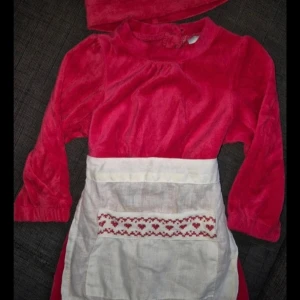 Jättefin tomtemordress, nyskick storlek 74/80 cl - Superfin röd sammetsklänning från Kappahl med lång ärm och vitt förkläde med broderade röda hjärtan. Klänningen har knytband i midjan och kommer med matchande röd luva med tofs. Perfekt för julens alla tillfällen. Storlek 74/80 cl.                                                Klänningen går även använda till andra tillfällen.             3 delad.                                                                           Som ny! Djur och rökfritt hem 