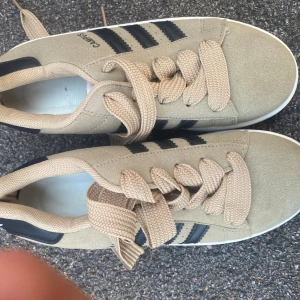 Beige Adidas Campus sneakers - Adidas Campus sneakers i beige mocka med klassiska svarta ränder på sidorna och svart häl med vit logga. Skorna har rund tå, platt sula och breda beige snören. Snygg retrostil med gummisula i brunt och vitt. Perfekt för dig som gillar streetwear och klassisk design.