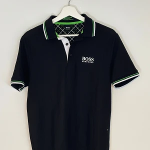 Svart Hugo Boss pikétröja - Snygg svart pikétröja från Hugo Boss med vit logga på bröstet. Kragen och ärmsluten har vita och gröna ränder som ger en sportig touch. Klassisk passform och korta ärmar, perfekt för en clean och stilren look.