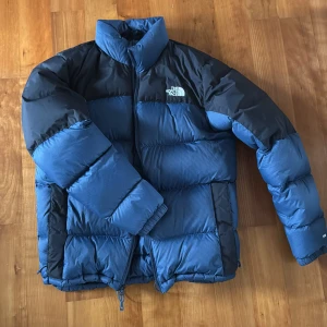 Blå dunjacka från The North Face - Säljer en blå och svart dunjacka från The North Face med hög krage och dragkedja framtill. Jackan har puffad design, justerbar nederkant med snörning och logga på bröstet och ryggen. Perfekt för kalla dagar och har en riktigt schysst vinterjacka. Nypris:3700kr