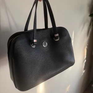 Svart handväska från Tommy Hilfiger - Snygg svart handväska från Tommy Hilfiger i strukturerat skinn med silverfärgade metalldetaljer och logotyp framtill. Väskan har ett innerfack som är perfekt för lite mindre grejer. 