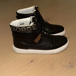 Svarta sneakers från Björn Borg - Snygga svarta sneakers från Björn Borg med högt skaft och tydlig logga i silver på hälen. Skorna har snörning, vit platt sula och är tillverkade i strukturerat skinn. Insidan är vit och de har en rund tå. Perfekta för dig som gillar streetstyle.