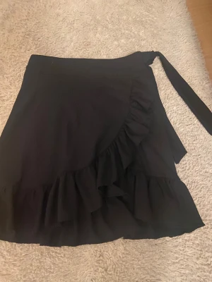 Svart omlottkjol med volang från Gina Tricot - Svart kort omlottkjol från Gina Tricot med snygga volangdetaljer längs kanten och knytband i midjan. Kjolen är gjord i mjukt polyestermaterial med lite stretch för skön passform. Perfekt för dig som gillar trendiga och feminina plagg.