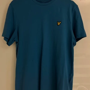 Blå t-shirt från Lyle & Scott - Snygg blå t-shirt från Lyle & Scott i mjuk bomull. Klassisk rund hals och korta ärmar. På bröstet sitter den ikoniska broderade gula fågeln som ger en stilren touch. Perfekt för dig som gillar enkel och clean design.