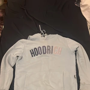 Ljusblå hoodie från Hoodrich XS - Säljer en ljusblå hoodie från Hoodrich i storlek XS. Tröjan har broderad HOODRICH-text i mörkblått och silver på bröstet, snörning vid huvan och en stor magficka. Perfekt för dig som gillar streetwear och vill ha en chill look.