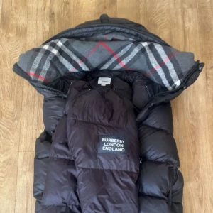 Svart pufferjacka från Burberry - Svart pufferjacka från Burberry London England med klassiskt rutigt foder i huvan. Jackan har dragkedja och knappar framtill, samt en stor huva med grått, vitt och rött rutmönster. Perfekt för kalla dagar med sin tjocka och fluffiga design.