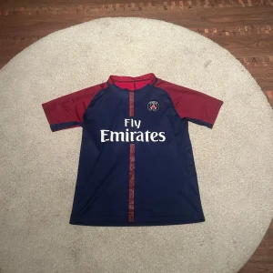 PSG Neymar Jr #10 fotbollströja - Säljer en officiell Paris Saint-Germain fotbollströja med Neymar Jr och nummer 10 på ryggen. Tröjan är marinblå med röda ärmar och detaljer, har korta ärmar och är tillverkad i lätt polyester. PSG-logga och Fly Emirates tryck på framsidan.