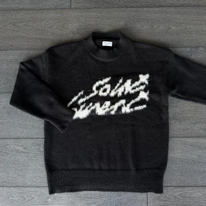 Svart stickad Saint Laurent tröja - Svart stickad tröja från Saint Laurent med vit, fluffig logotyp framtill. Tröjan har rund hals, långa ärmar och ribbade muddar. Perfekt för dig som vill ha en statement piece med lyxig känsla och cool design.