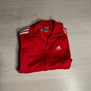 Röd Adidas zip - Snygg röd Adidas tröja med klassiska vita ränder längs ärmarna och logga på bröstet. Tröjan har dragkedja framtill, ribbade muddar och krage samt två fickor med dragkedja. Perfekt för dig som gillar sportig stil och streetwear.