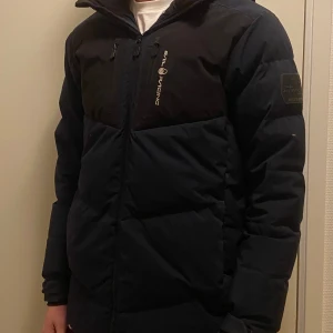 Vinterjacka från Sail Racing - Säljer en mörkblå pufferjacka från Sail Racing med huva, två bröstfickor med dragkedja och två vanliga fickor. Perfekt för vintern. Jackan har en liten fläck i bak som säkrt går att ta bort (har ej provat). Storlek. Är 176 men är som en S Hör av er vid frågor eller fler bilder. 