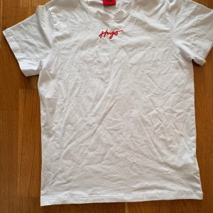 Vit Hugo Boss t-shirt med röd logga - Vit t-shirt från Hugo Boss med röd broderad Hugo-logga på bröstet. Klassisk rund halsringning och korta ärmar. Tillverkad i mjuk bomull för en skön känsla. Perfekt för en clean och stilren look.