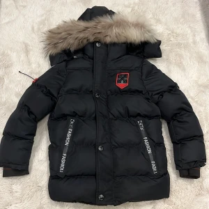 Svart pufferjacka Off-White med päls - Svart pufferjacka från Off-White med fet päls på huvan och coola logopatchar i rött och svart. Jackan har dragkedjor med texten 'FASHION', knappar framtill och snörning vid ärmarna. Insidan är mjuk och fluffig, perfekt för kalla dagar.