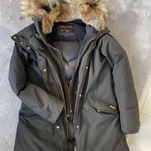 Svart parkas från Woolrich med päls - Svart lång parkas från Woolrich med huva och snygg pälsdetalj. Jackan har dragkedja och knappar framtill, två stora fickor och är fodrad för extra värme. Materialet är en mix av bomull och nylon, perfekt för kalla vinterdagar.