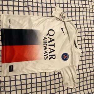 PSG Mbappé vit matchtröja Nike - Säljer en officiell Paris Saint-Germain fotbollströja med Mbappé och nummer 7 på ryggen. Tröjan är vit med ett vertikalt fält i blått och rött framtill, PSG-logga på bröstet och Nike Swoosh. Materialet är lätt polyester, perfekt för träning eller match.