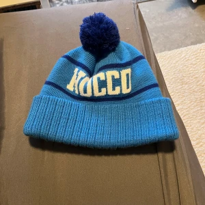 Blå NOCCO mössa med tofs - Snygg blå mössa från NOCCO med mörkblå ränder och stor tofs på toppen. Stickad design med uppvikt kant och vit NOCCO-logga framtill. Perfekt för kalla dagar och ger en sportig vibe till din outfit.