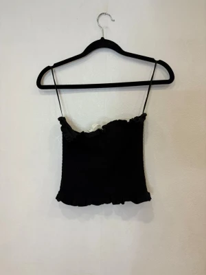 Svart smockad linnetopp med volang - Supersöt svart linnetopp med smockad struktur och tunna axelband. Toppen har volangdetaljer både upptill och nedtill som ger en gullig touch. Perfekt att styla med jeans eller kjol för en trendig look.