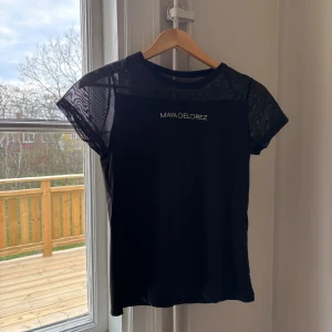 Svart mesh-topp från Maya Delorez - Snygg svart topp från Maya Delorez med meshpartier över axlar och ärmar. T-shirten har rund halsringning och korta ärmar, samt diskret logga framtill. Perfekt för en sportig och trendig look.