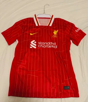 Liverpool Salah #11 matchtröja Nike - Liverpool FCs officiella matchtröja med M. Salah #11 på ryggen. Röd med gula och vita detaljer, tunna gula ränder och klubbmärke broderat på bröstet. Tillverkad i lätt och ventilerande polyester, kortärmad och med klassisk krage. EJ ORIGINAL!